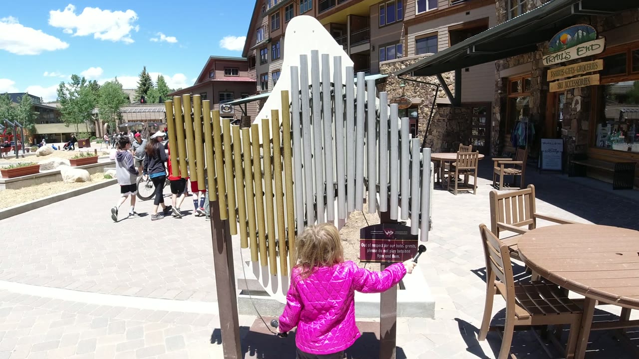 The girl raises a mallet or striker toward the white chimes in Winter Park, Colorado, with a shopfront, benches, and passing visitors behind her.