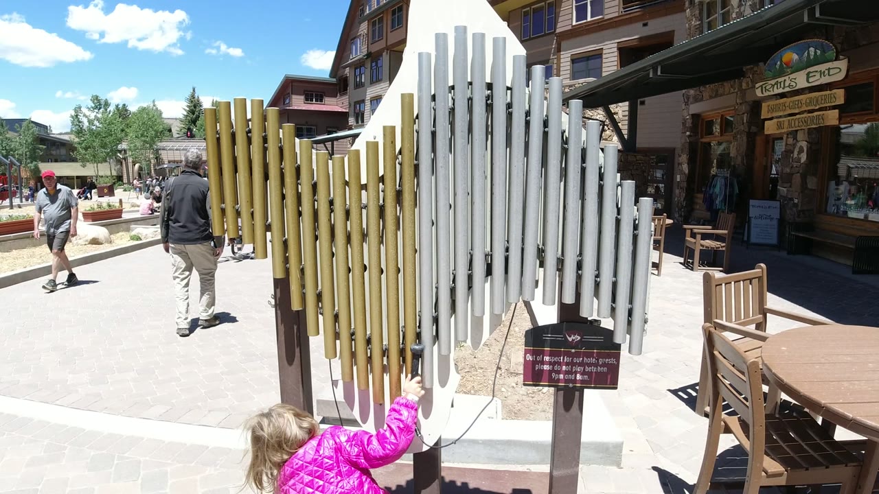 Child in a pink coat reaches toward outdoor musical pipes beside a walkway.