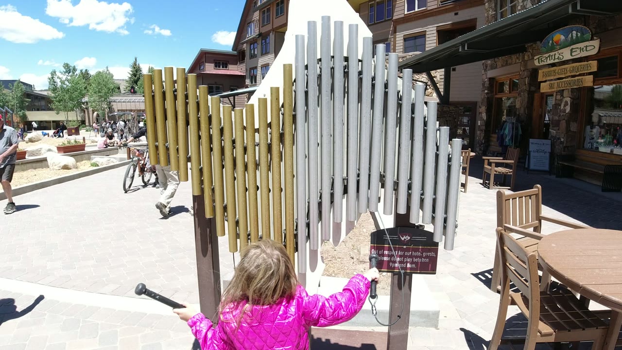 The child stands closer to the vertical pipes in Winter Park, Colorado, while a cyclist and pedestrians pass through the sunlit walkway behind the instrument.