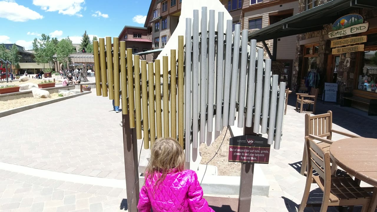 Child in a pink jacket stands behind outdoor chimes in a sunny plaza.