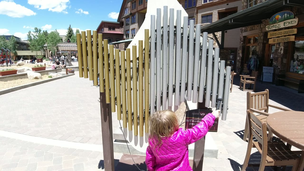 The child reaches toward the white chime tubes in Winter Park, Colorado, with a wooden storefront, patio table, chair, and directional-style sign in the background.