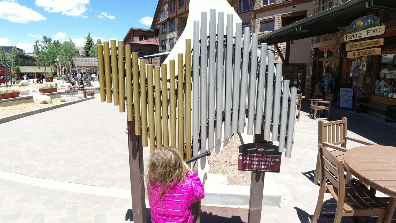 At 0:00 in Winter Park, Colorado, a young girl in a pink jacket stands with her back to camera and reaches toward yellow and white outdoor chime tubes in a sunny plaza.