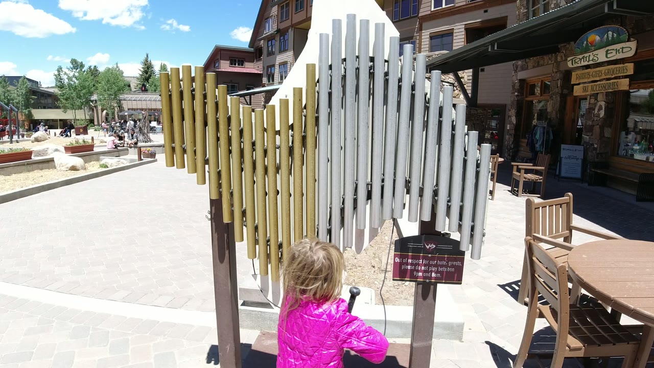 At 0:01 in Winter Park, Colorado, the girl has shifted slightly right while facing the white chime tubes, with a small sign, wooden bench, storefront, and open plaza behind her.