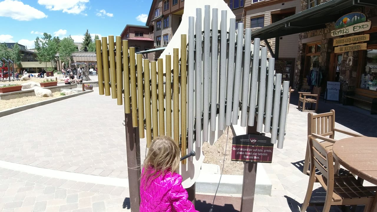 Child in a pink jacket beside large outdoor chimes in a sunny plaza.