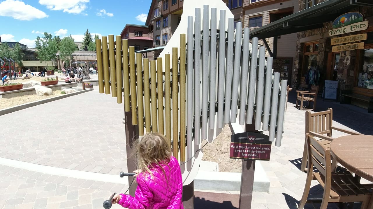 The child shifts closer to the chime structure in Winter Park, Colorado, holding a mallet near the tubes while storefronts, benches, and people remain in the background.