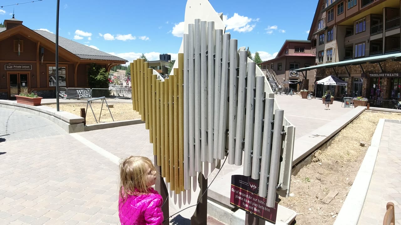 Child in a pink jacket beside a vertical tube sculpture in a sunny plaza.
