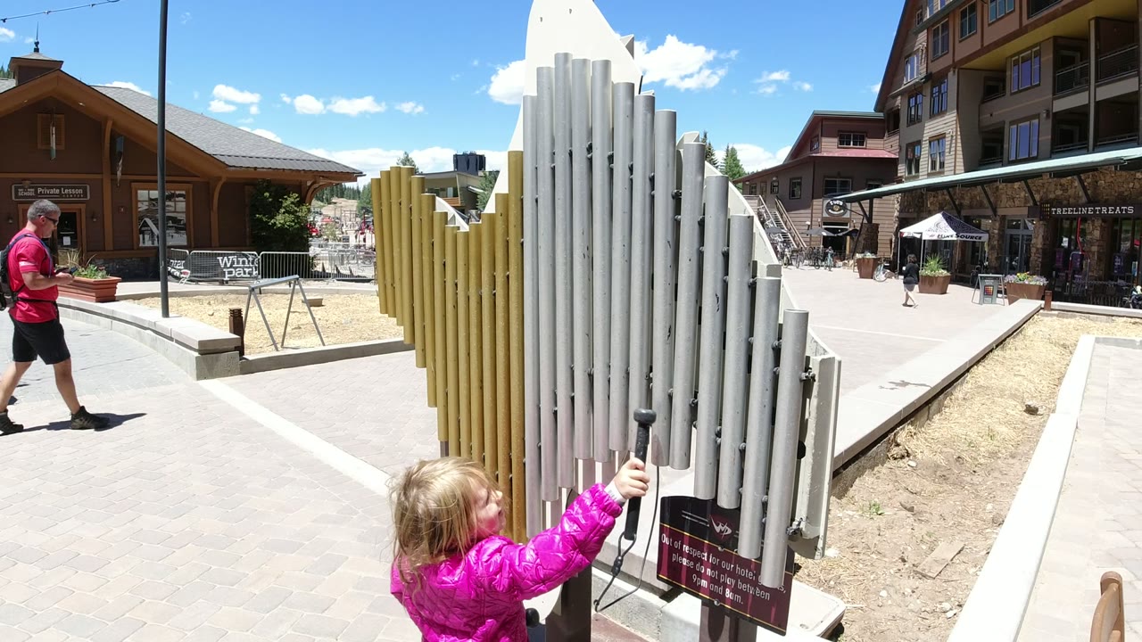 The child extends a stick-like object toward the white and yellow tube sculpture in Winter Park, Colorado, while a man in a pink shirt walks through the plaza behind it.