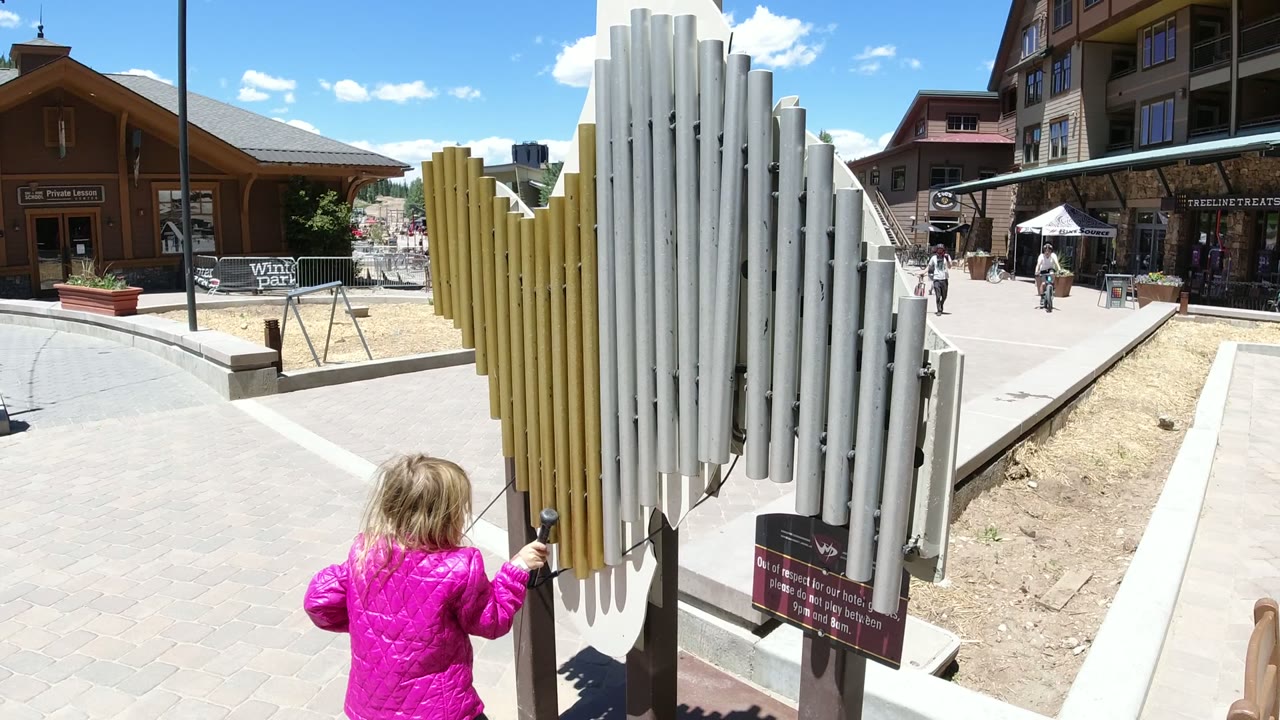 Blonde child in a pink jacket reaches toward outdoor chimes in a pedestrian plaza.