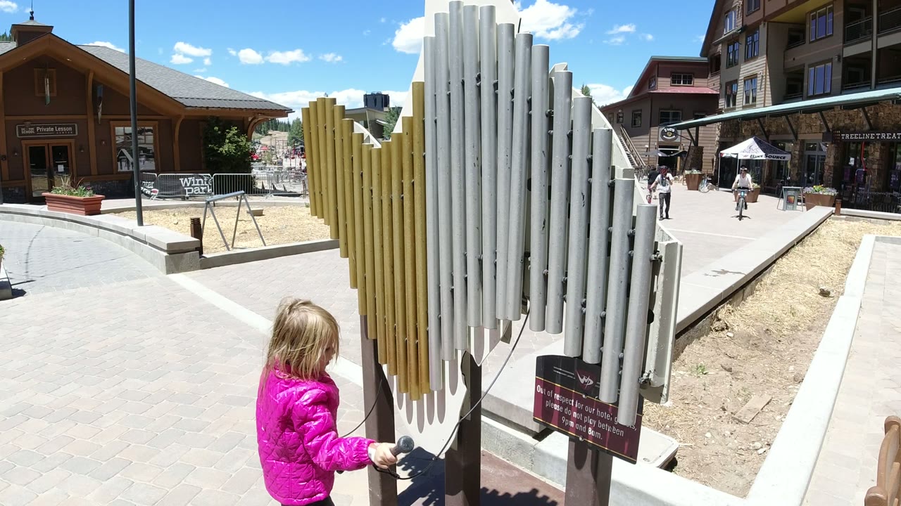 The child shifts beside the yellow and silver chime tubes in Winter Park, Colorado, while a paved plaza, shopfronts, and distant people remain in view.