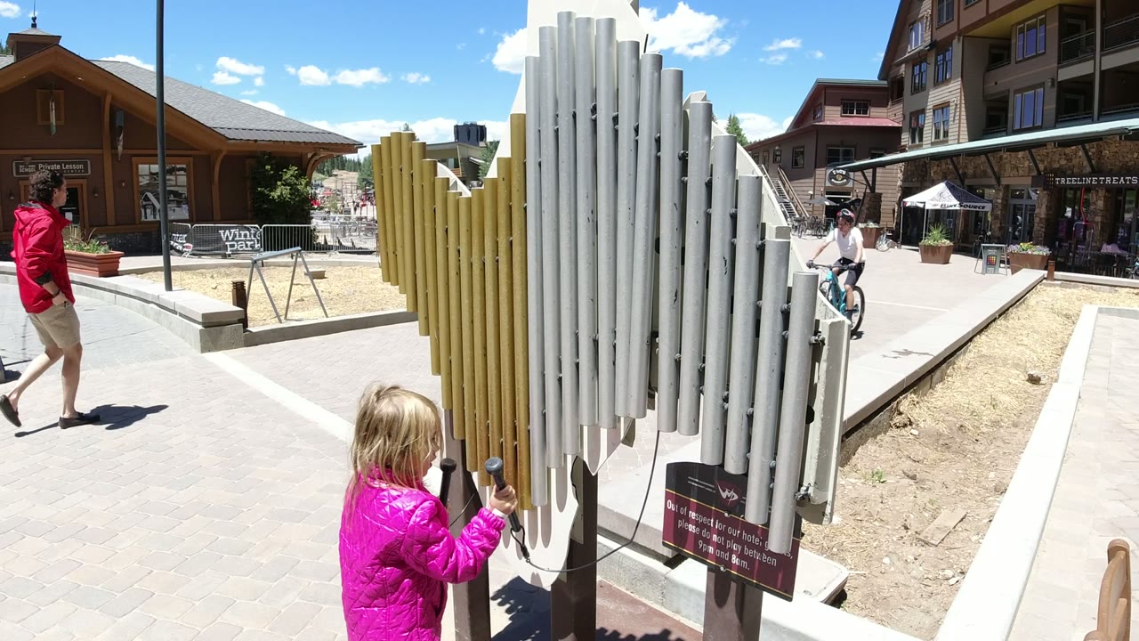A blonde child in a bright pink jacket strikes a large outdoor chime instrument in Winter Park, Colorado, with storefronts and a passing adult in the background.