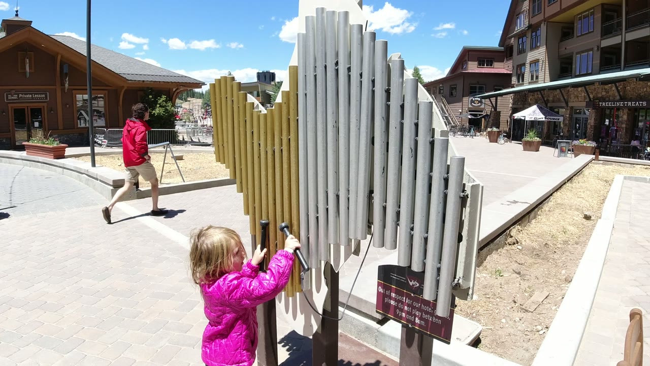 The child in the pink jacket reaches higher on the chime bars in Winter Park, Colorado, while an adult walks across the sunny plaza behind the instrument.