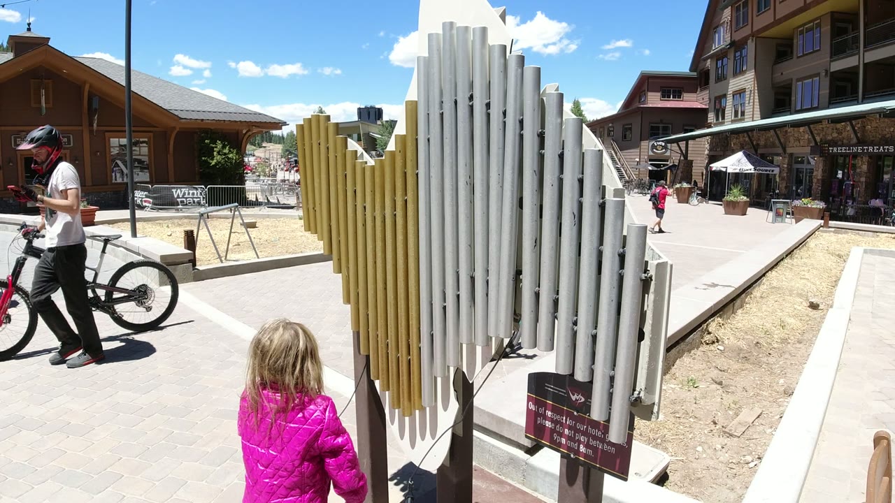 Child in a pink jacket near outdoor chimes in a sunny plaza.