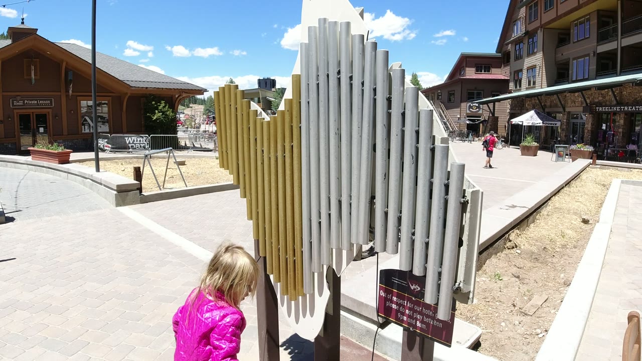 The white and yellow chime structure fills the foreground in Winter Park, Colorado, while the child in pink is partly cropped at the lower edge and pedestrians cross the plaza beyond.