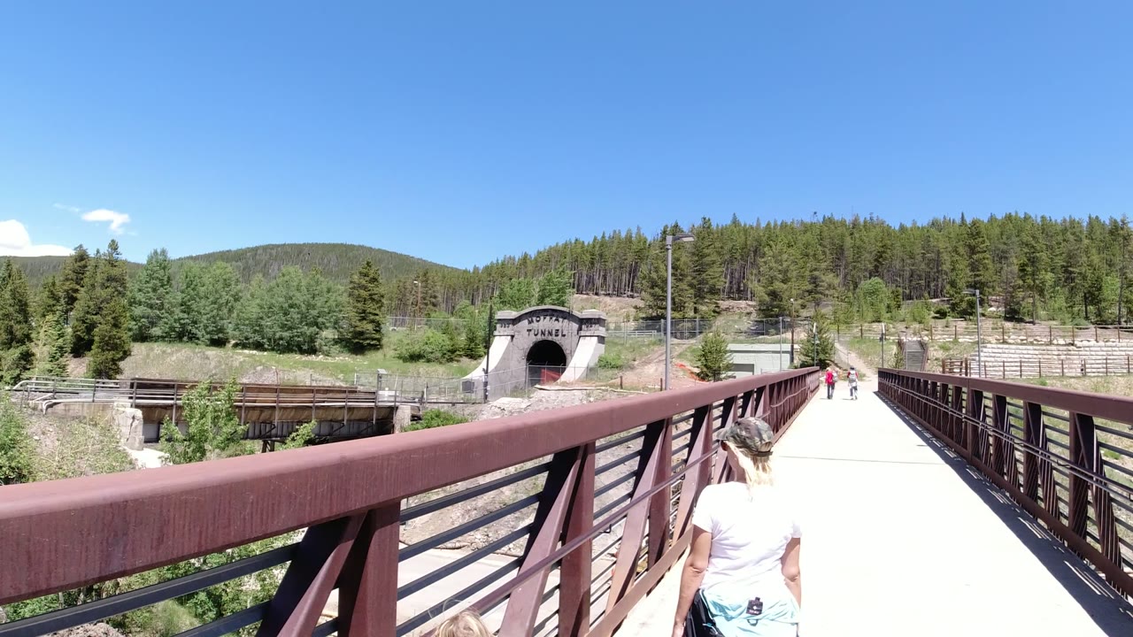 A woman seen from behind walks along a red-brown pedestrian bridge in Winter Park, Colorado, with a tunnel portal ahead, forested hills, and bright blue sky.