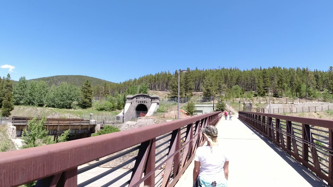 The bridge railings lead toward the tunnel entrance in Winter Park, Colorado, while the woman remains near the lower edge and small pedestrians appear farther ahead.