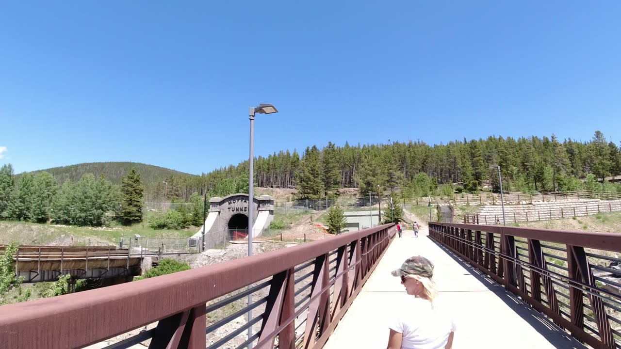 Wide view along a red-railed bridge toward a tunnel entrance in Winter Park, Colorado, with pine-covered hills, blue sky, and a partially visible person at the lower foreground.