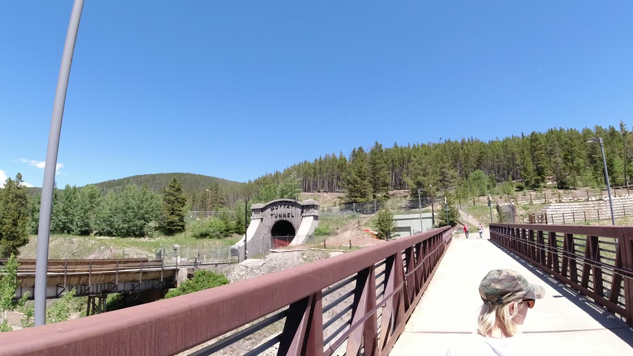 The Winter Park bridge remains centered, with the tunnel entrance ahead, red-brown railing across the foreground, trees and hillside in back, and a woman partly visible at lower right.