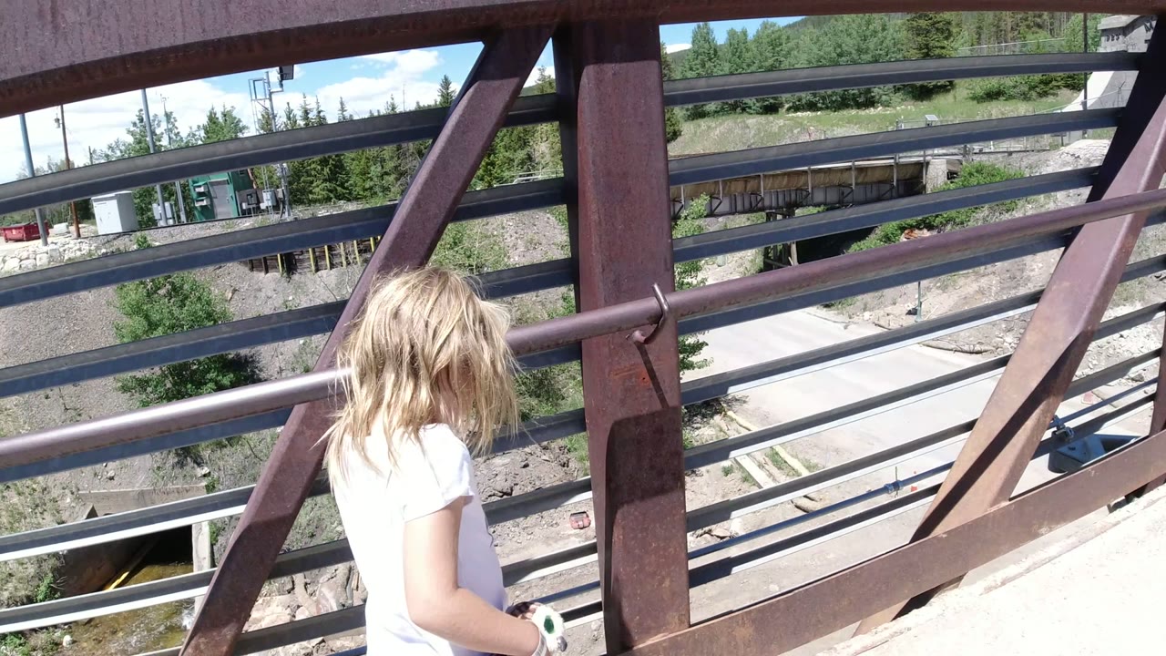 A blonde child in a white shirt walks along a metal pedestrian bridge in Winter Park, Colorado, with road and trees visible through the railings.