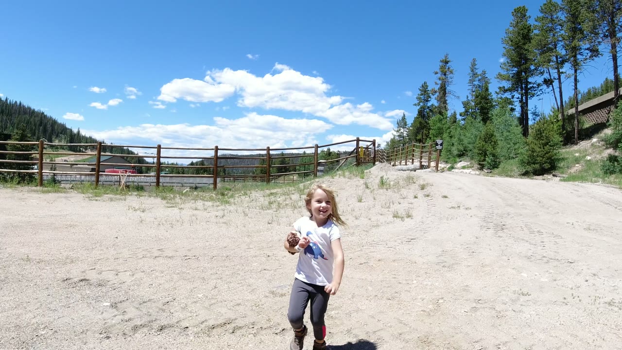 A young girl in Winter Park, Colorado holds a small object while standing on sandy ground, with a wooden fence, pine trees, hills, and blue sky behind her.