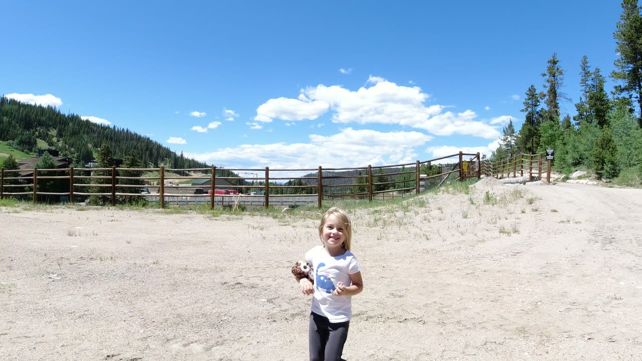A young girl in Winter Park, Colorado stands near the center of a wide sandy clearing, with a wooden rail fence, distant buildings, pine-covered slopes, and bright clouds in view.