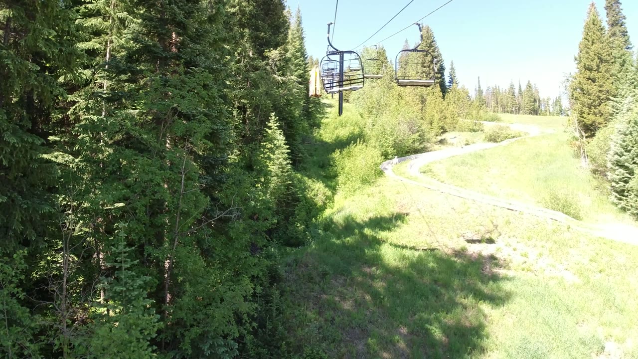 At 0:01 in Winter Park, Colorado, the chairlift appears slightly farther along the cable above conifers, with a pale curving path visible across the sunlit green slope.