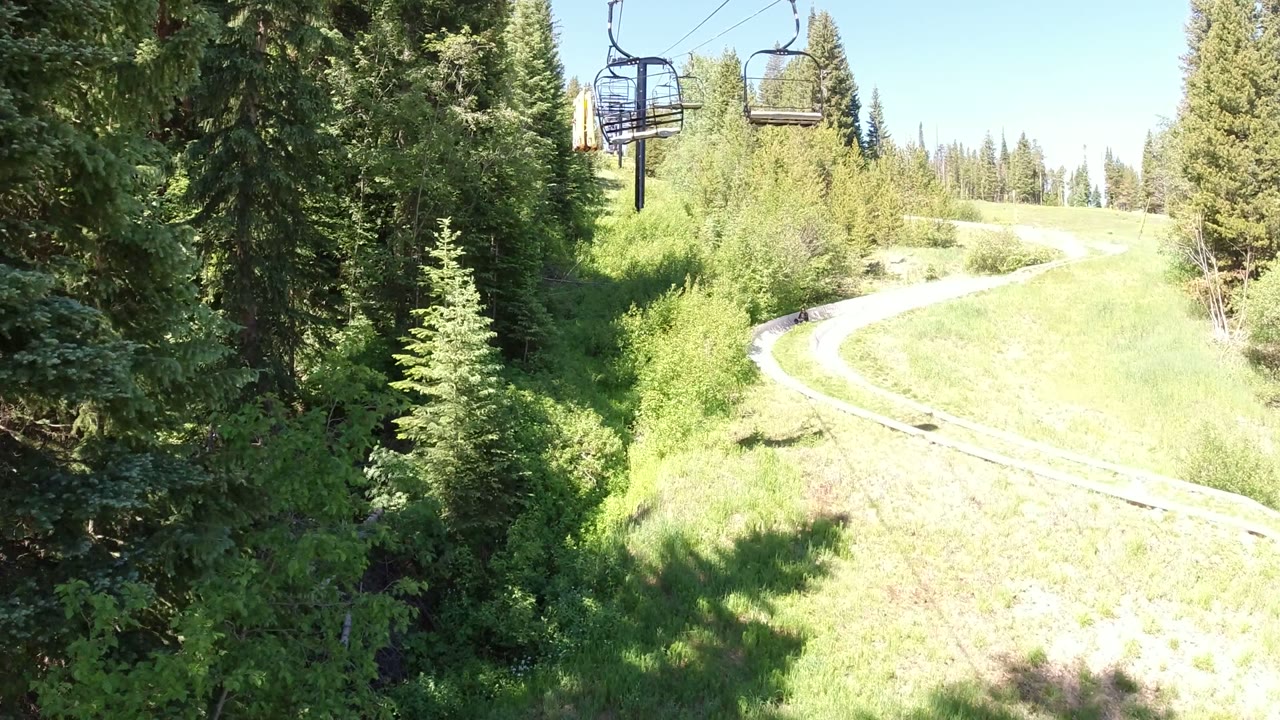 One second in, the empty lift chairs sit near the top of frame while evergreen trees frame a pale curved track or path across the sunny Winter Park slope, with no visible people.