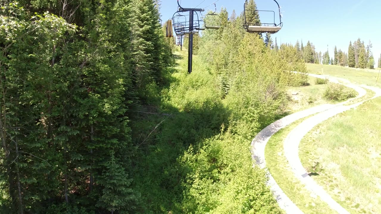 Opening frame in Winter Park, Colorado with empty chairlift seats overhead, a pale winding path below, dense green trees, bright sun, and no visible people.