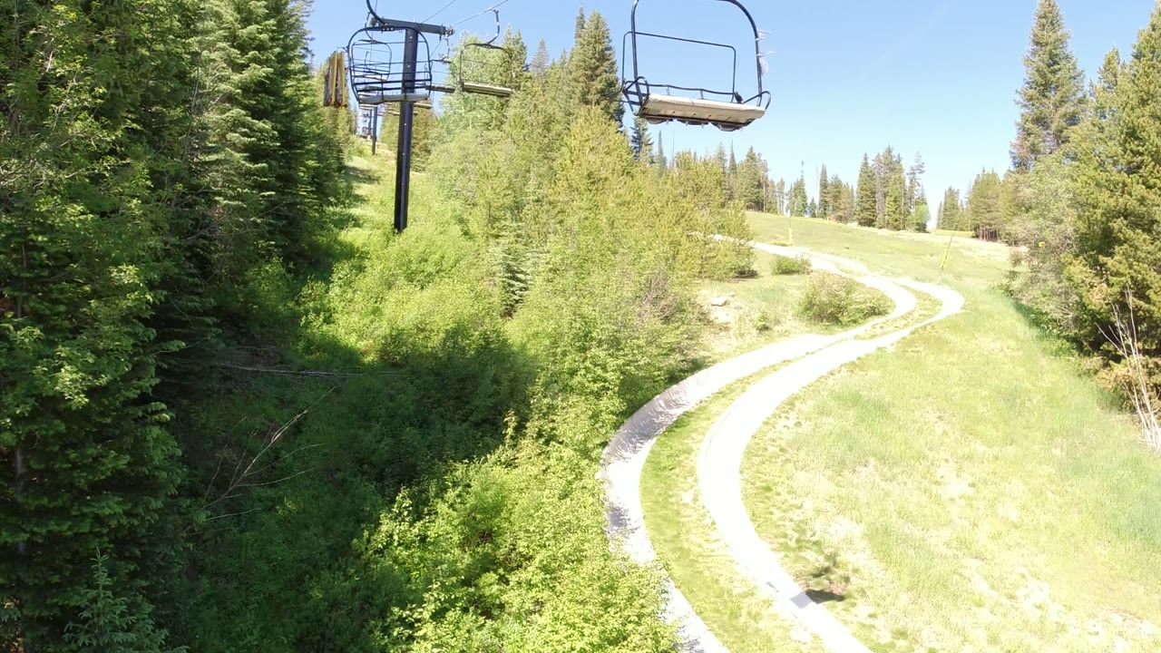 Winter Park, Colorado view with chairlift chairs near the top of frame, a sunlit grassy slope, thick evergreens, and the path bending uphill with no visible people.