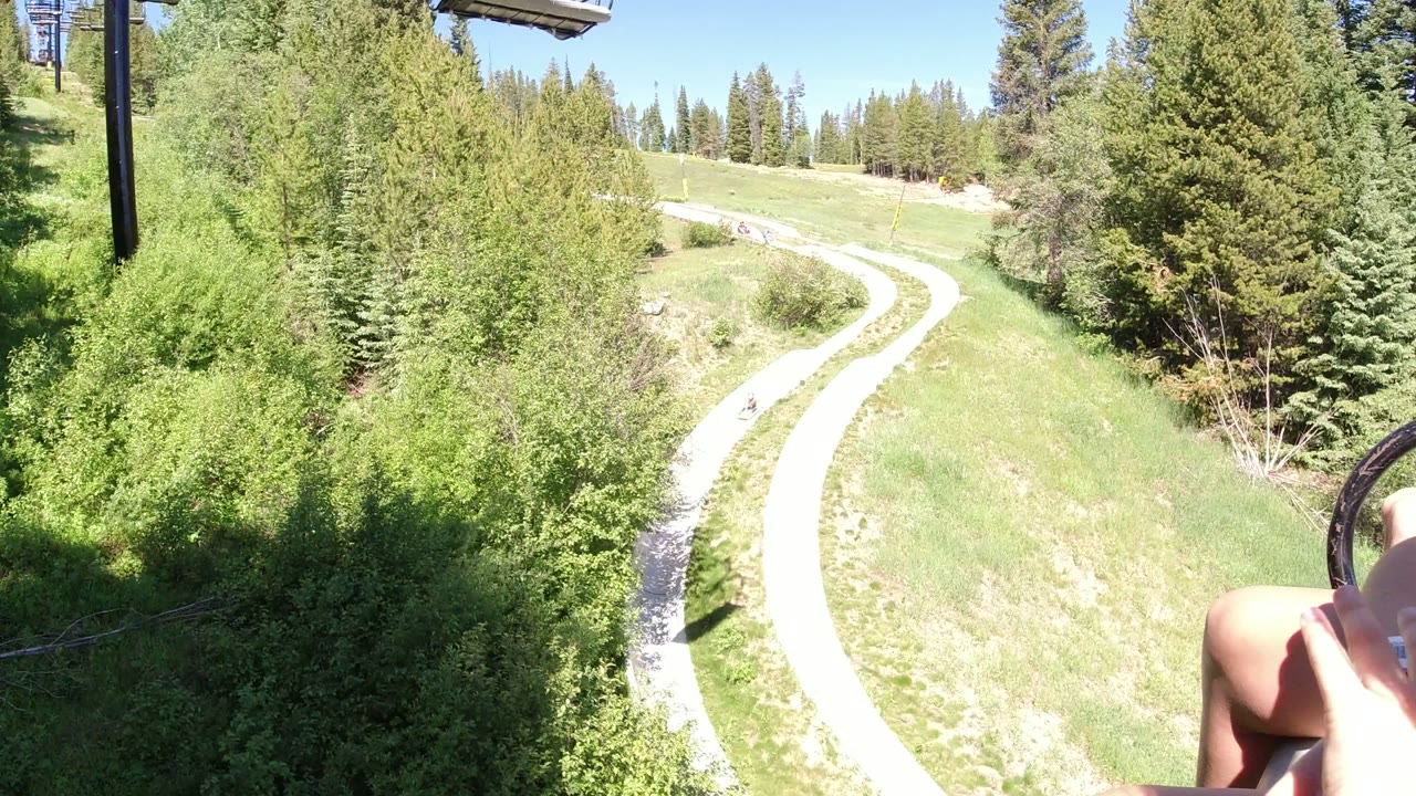 The Winter Park forest path curves through sunlit grass while a rider's bare legs and lift bar appear along the right side of the frame.