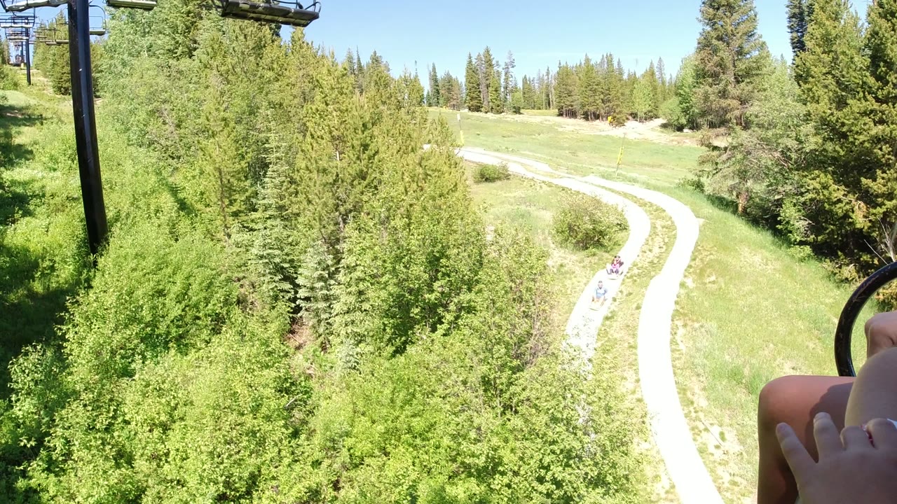Winter Park, Colorado chairlift view over dense green trees, a pale curving road or trail, blue sky, and the rider's legs at the lower right edge.