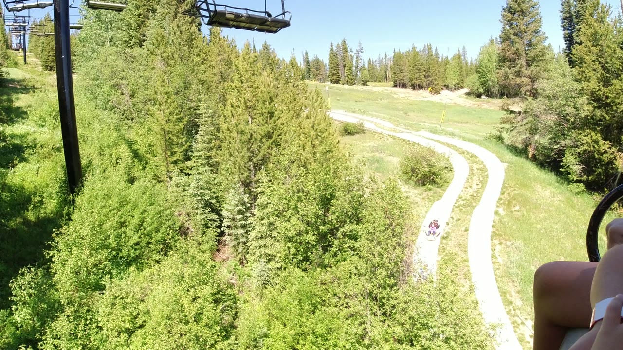 Winter Park, Colorado lift POV showing bright evergreen forest, a curving service road or trail, overhead chairlift hardware, and a rider's hand near the right edge.