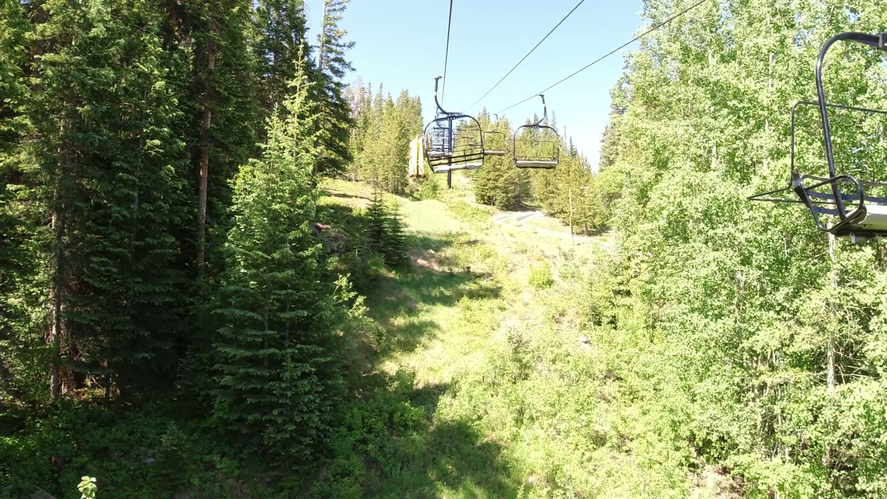 Empty chairlift seats hang above a grassy slope and dense trees in Winter Park, Colorado, with no visible people at the opening frame.