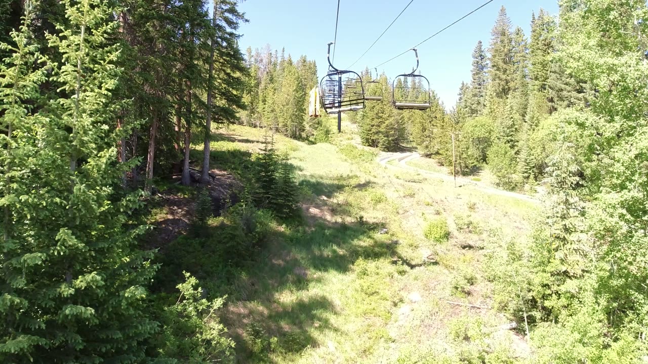 A chairlift chair hangs near the upper right while the cable line leads across a bright grassy slope and evergreen forest in Winter Park, Colorado, with no visible people.