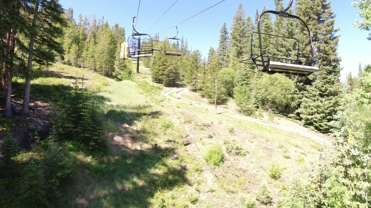 An elevated Winter Park, Colorado view shows chairlift cables crossing above a grassy slope, evergreen trees, a winding path, and no visible people.