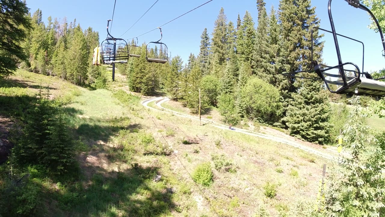 An empty lift chair appears near the upper right over the Winter Park hillside, with pine trees, a pale winding path, and no visible people below.