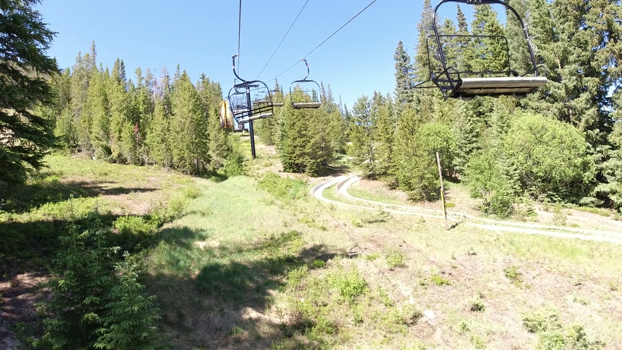 Chairlift seats above a sunny forested hillside and winding dirt road.