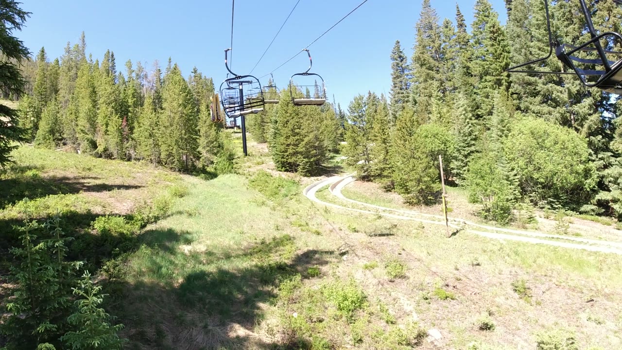 Second frame shows the chairlift line slightly farther along the forested Winter Park slope, with empty-looking seats overhead and the road curving through the grass below.