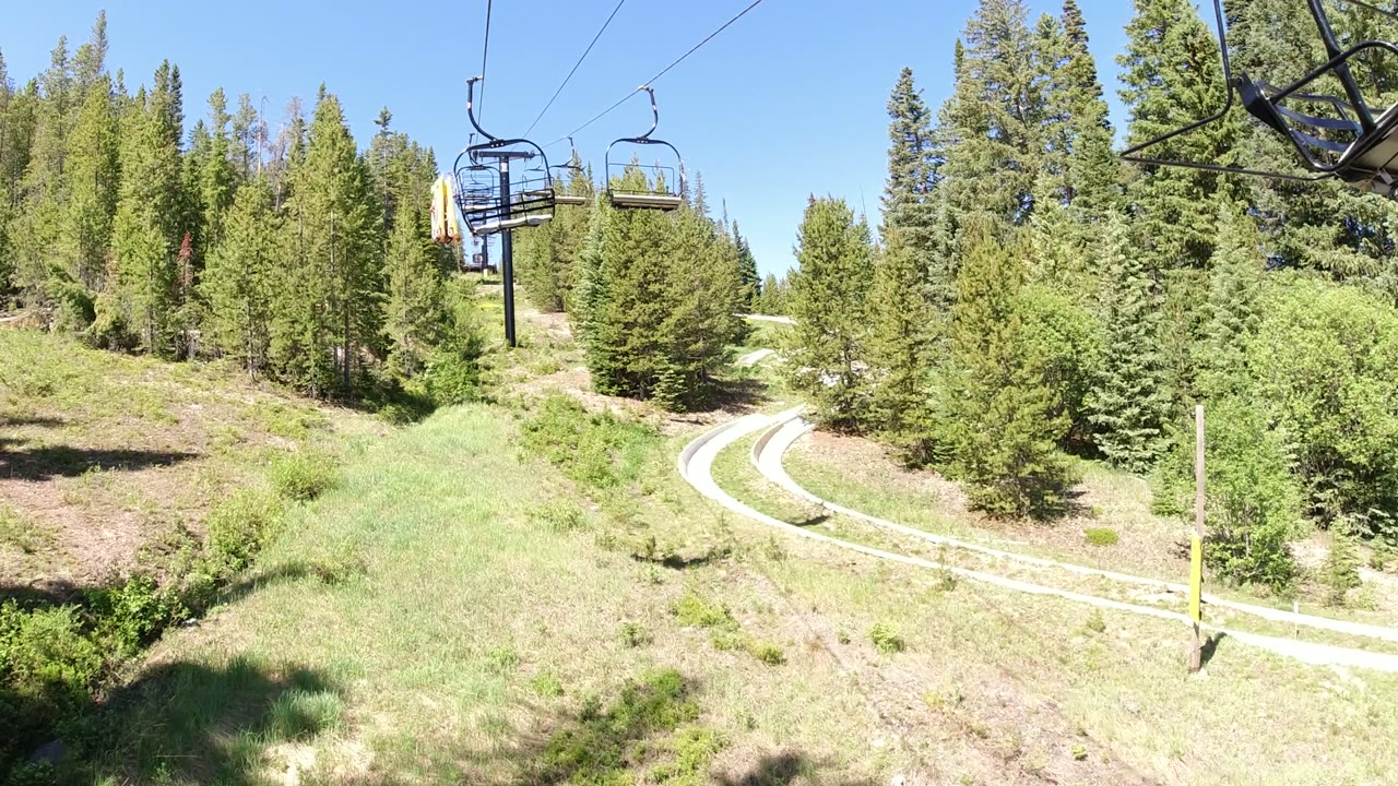 Winter Park, Colorado hillside view with empty lift chairs nearer the upper frame, dense green trees across the slope, a pale track curving through the lower scene, and no visible people.