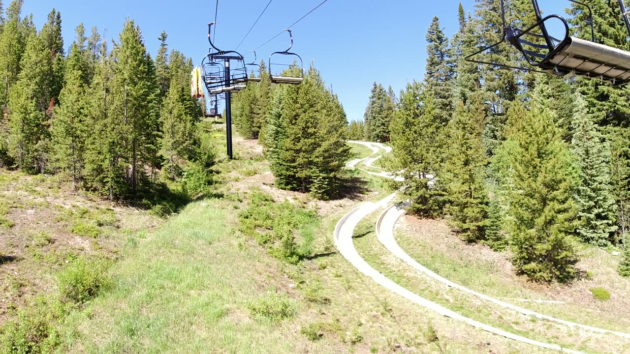 The Winter Park, Colorado view holds on chairlift seats above dense conifers, with the light-colored track curving from the lower right into the trees and no clearly visible people.