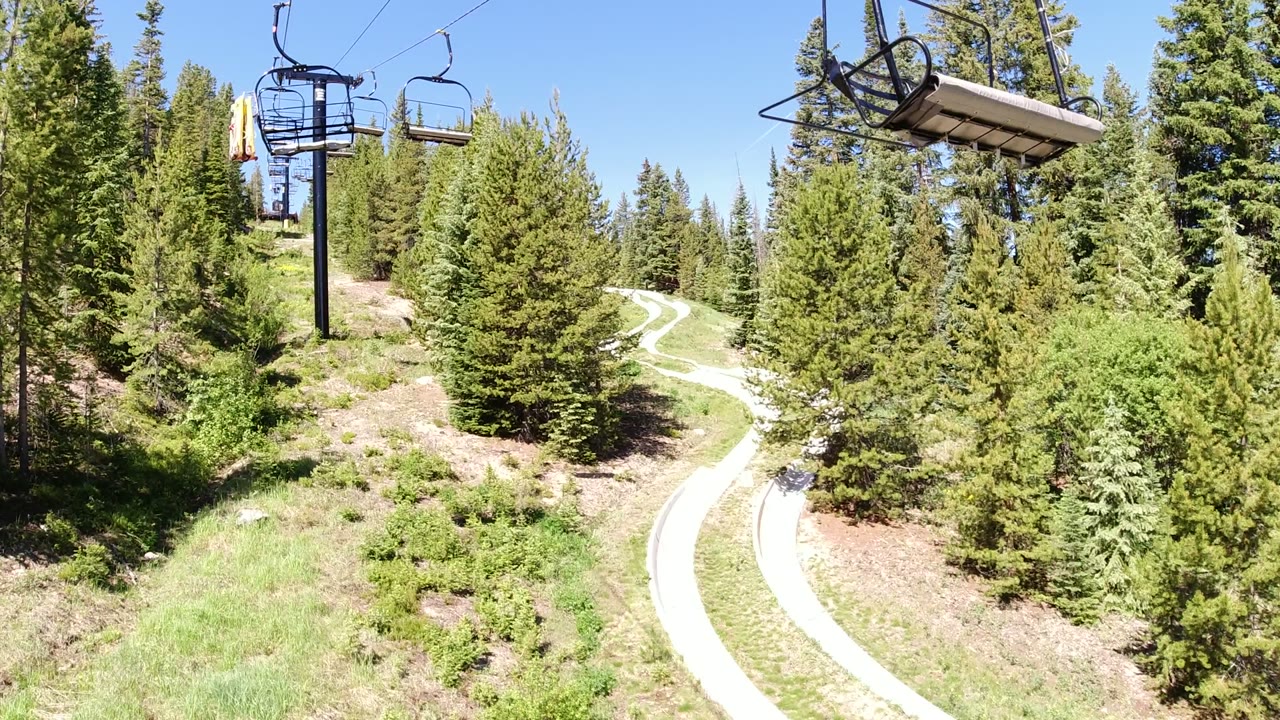Winter Park, Colorado frame with the chairlift line near the top edge, evergreen trees filling the slope, a winding pale path in the lower center, and no clearly recognizable people visible.