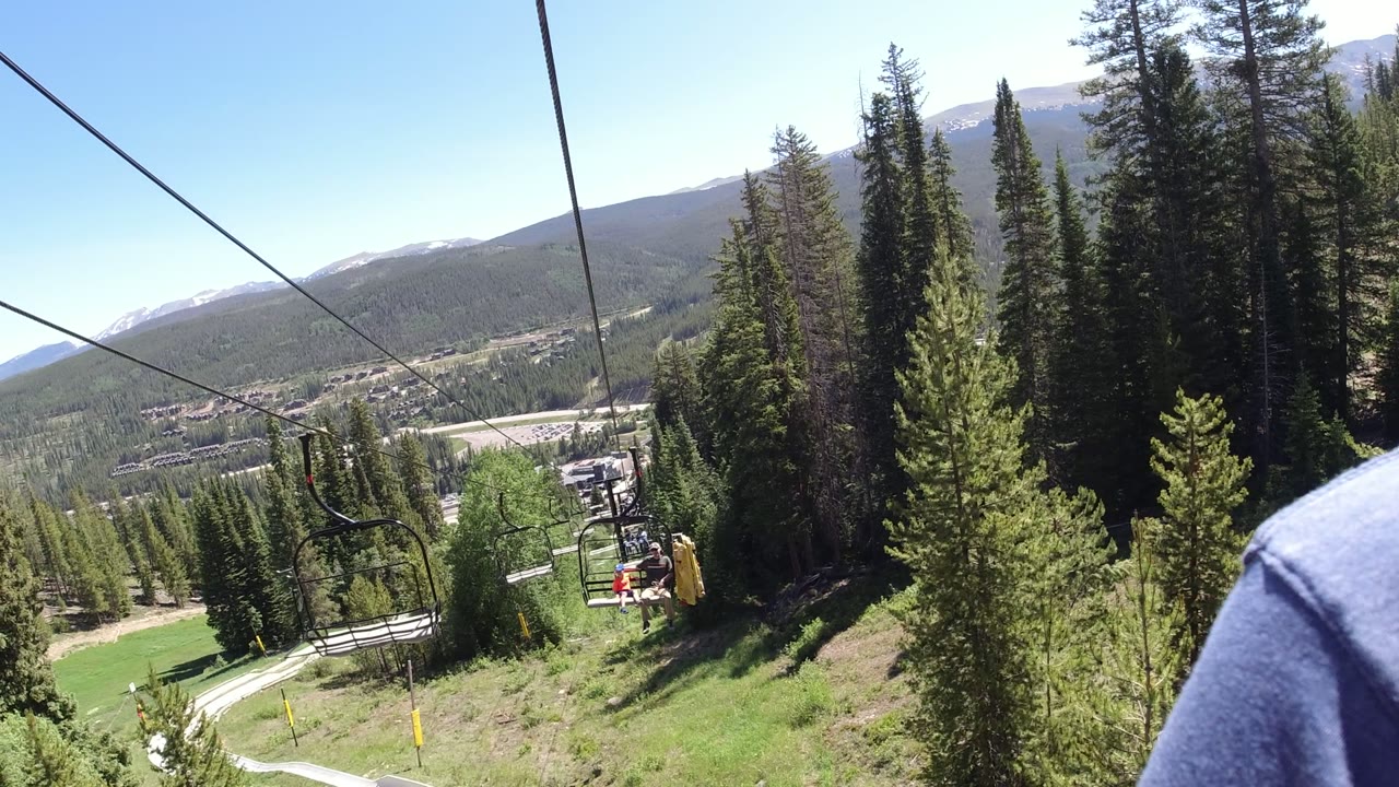 Opening view from a Winter Park, Colorado lift line with chairlift cables overhead, evergreen trees on the slope, valley buildings below, distant mountains, and no recognizable people visible.