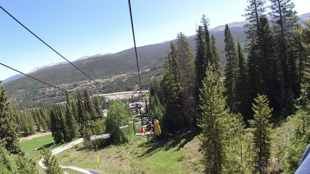 A Winter Park, Colorado chairlift line runs down the wooded slope toward valley buildings, with tall evergreens at right, mountains on the horizon, and no recognizable people visible.