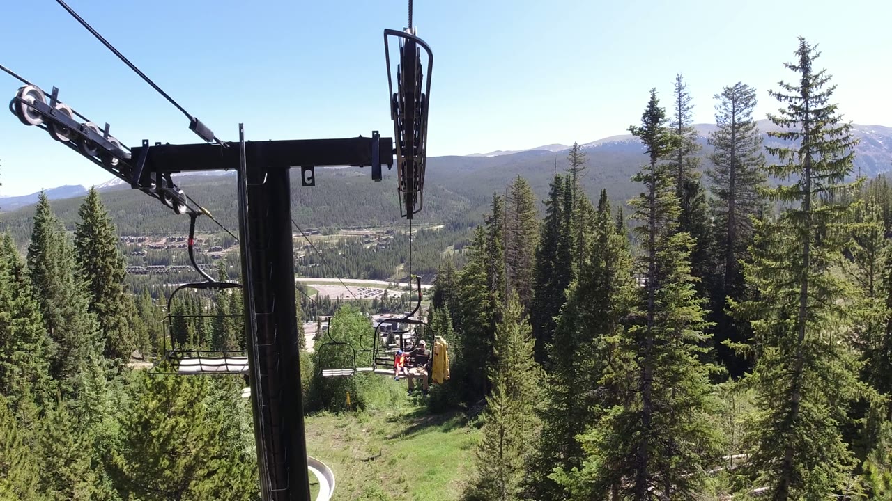Chairlift tower and cables above evergreen trees with mountains beyond.