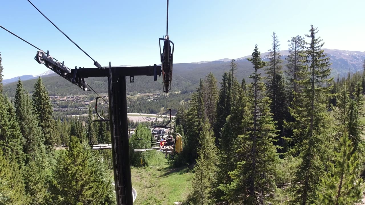 Winter Park, Colorado chairlift hardware fills the left foreground while cables lead toward small lift seats, green conifers, a valley floor, and pale mountain ridges, with no recognizable people visible.