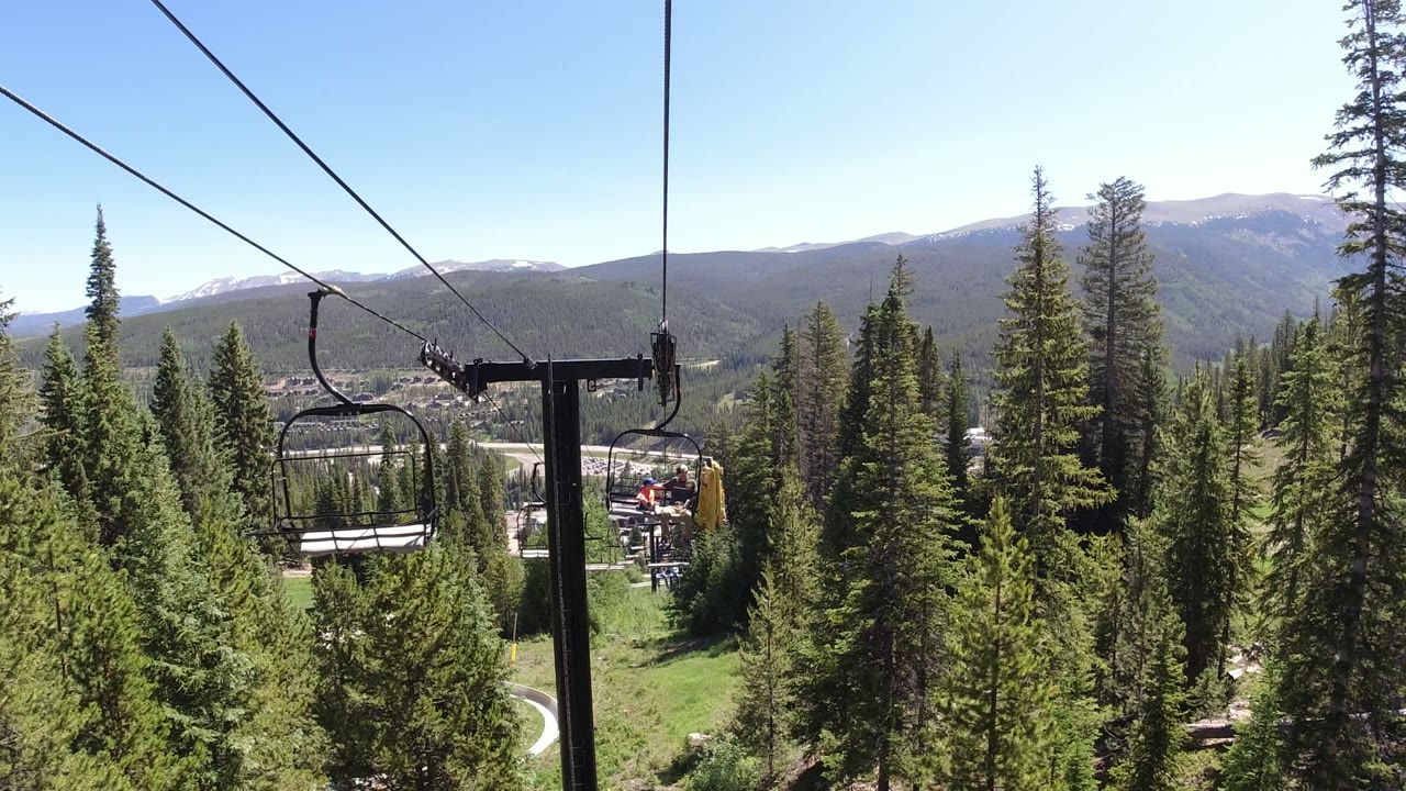 Opening view from a Winter Park, Colorado ropeway, with chairlift cables crossing above green pines, distant riders, and mountains under clear daylight.
