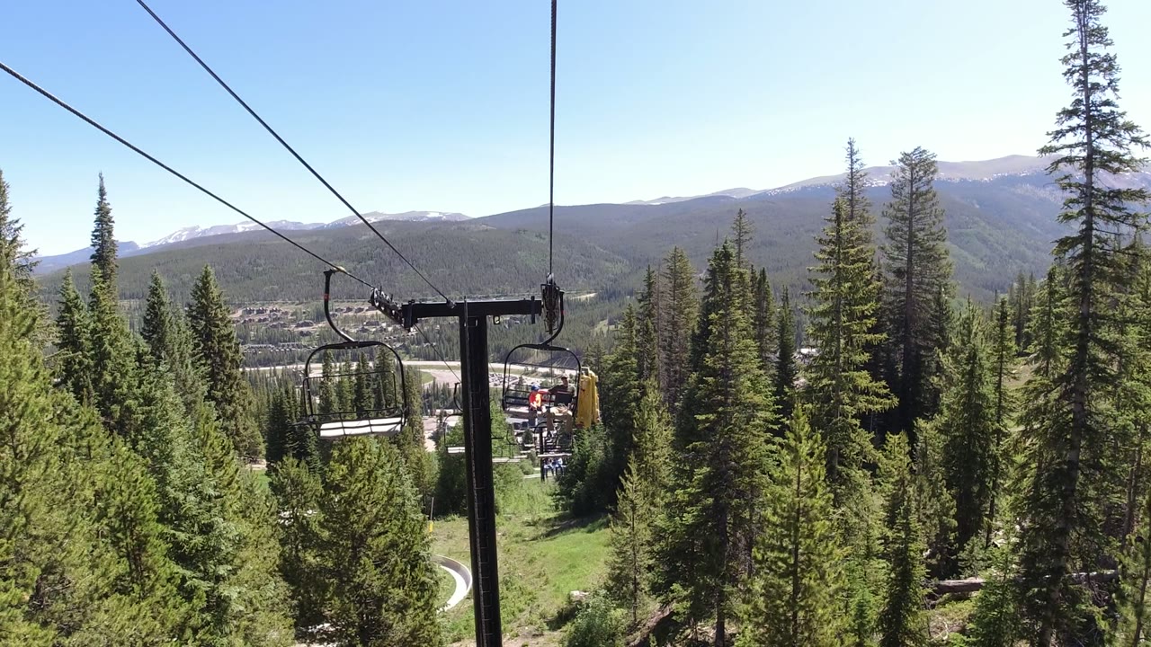 Winter Park, Colorado lift tower and chairlift seats sit near the center of frame, with evergreen forest below and a mountain ridge beyond.