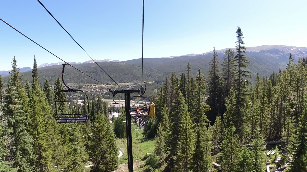 Opening frame of a Winter Park, Colorado chairlift view, with lift cables and empty chairs over dense evergreens, a valley, and distant mountains, with no visible people.