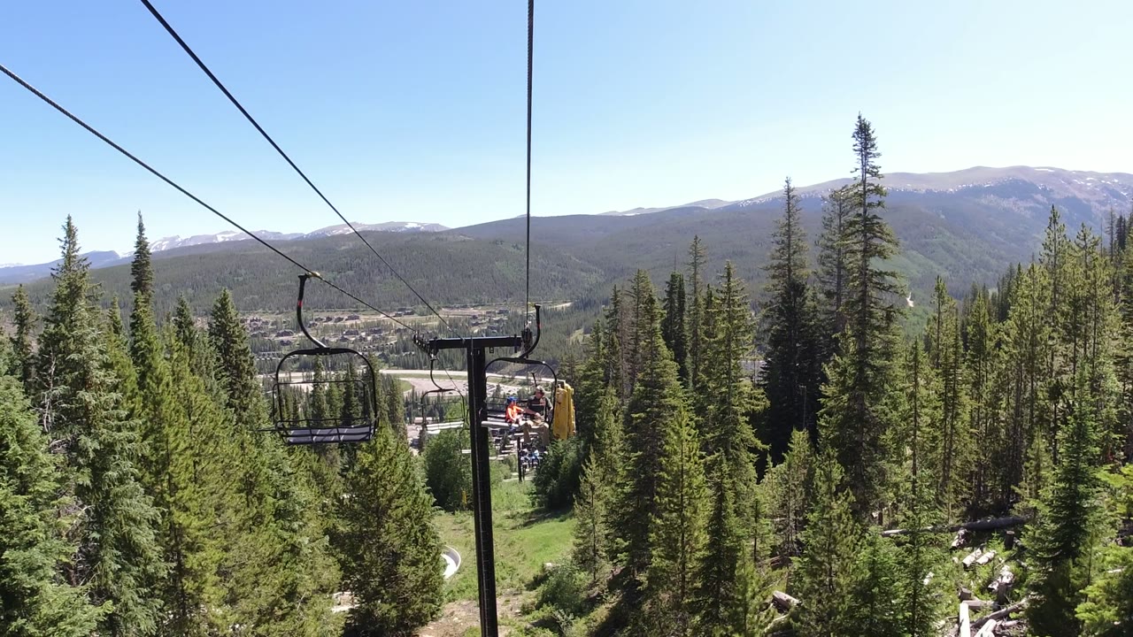 A Winter Park, Colorado lift tower sits near the center as chairlift cables run overhead above evergreen trees and a mountain ridge, with no recognizable people visible.