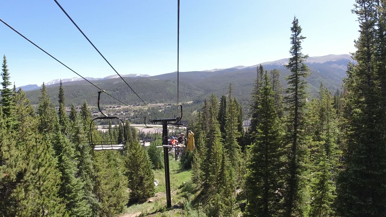 A chairlift in Winter Park, Colorado carries small distant riders past evergreen trees, with a sunlit valley and layered mountains in the background.