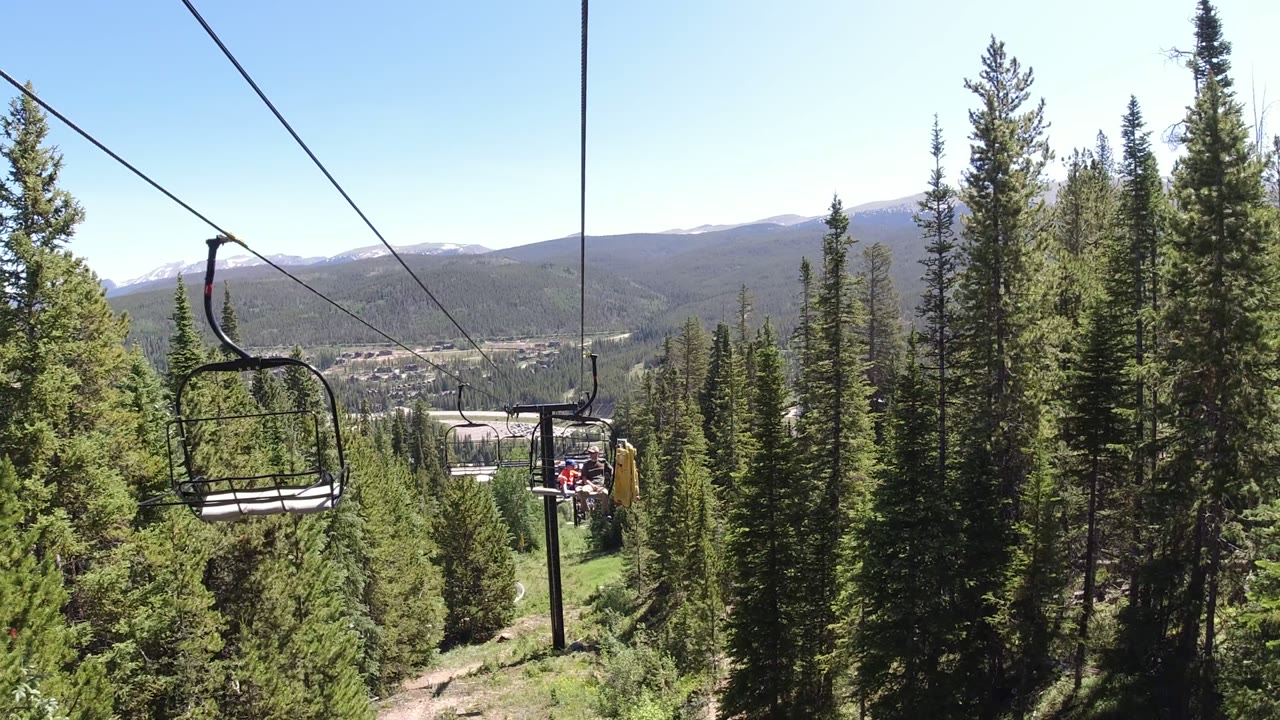 Opening view from a Winter Park, Colorado cableway, with empty chairlift seats at left, dense evergreen trees below, distant mountains, blue sky, and no visible people.